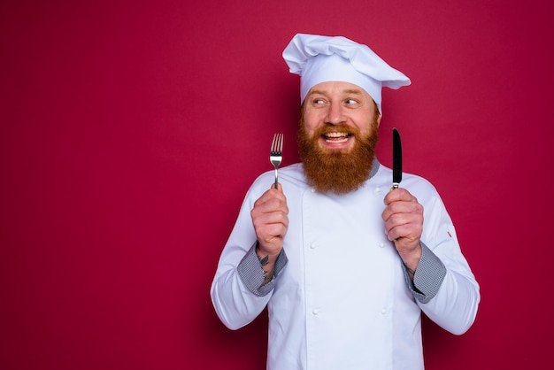 Feliz chef con barba y delantal rojo tiene cubiertos en la mano