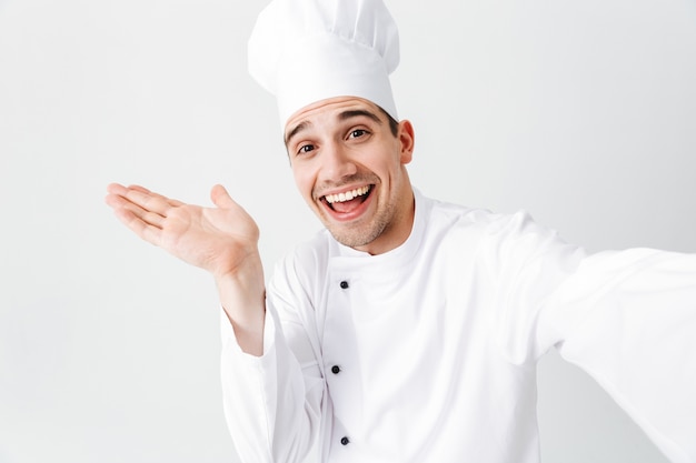 Feliz chef cozinheiro usando uniforme em pé, isolado na parede branca, tirando uma selfie