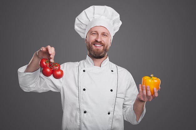 Feliz chef hombre barbudo mirando a la cámara con una sonrisa y demostrando tomates maduros y pimienta sobre fondo gris