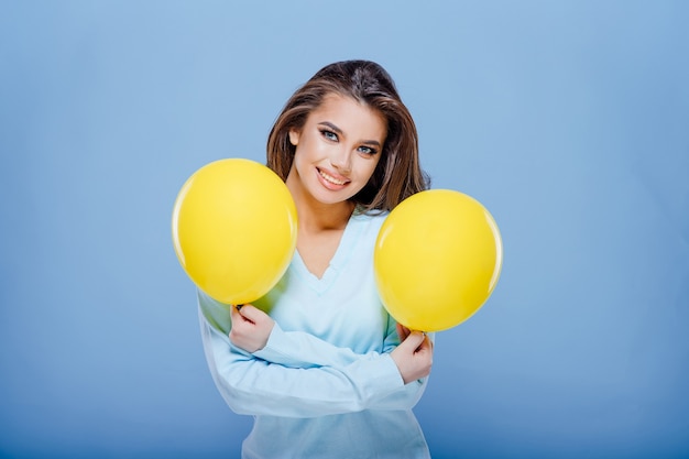 Feliz chica adolescente está sosteniendo dos globos amarillos y sonriendo para la cámara mientras se divierte contra i ...