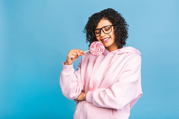 Feliz chica afro americana sexy comiendo piruletas. Mujer de modelo de glamour de belleza con caramelo dulce de piruleta de colores, aislado sobre fondo azul. Dulces