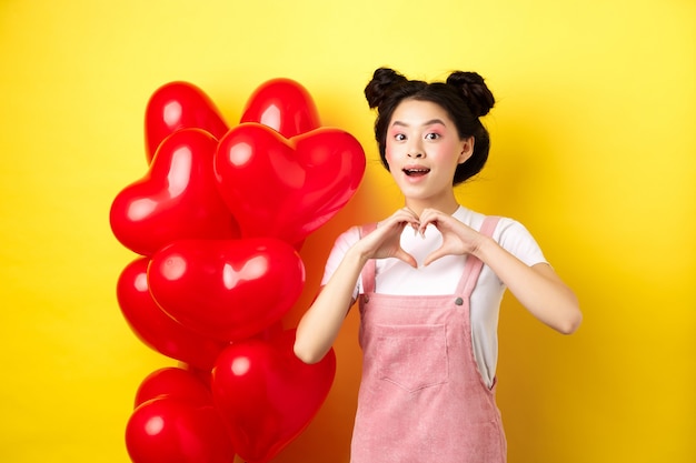 Feliz chica asiática mostrando signo de corazón cerca de globos románticos