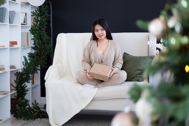 feliz chica asiática con ropa acogedora con caja de regalo sentada en el sofá en casa, tiempo de Navidad