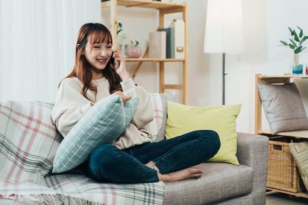 feliz chica asiática sentada con las piernas cruzadas en el sofá se ríe y sostiene una almohada mientras habla por teléfono con un amigo durante el día en una acogedora sala de estar en casa.