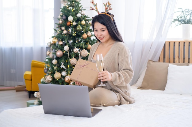 feliz chica asiática sosteniendo una copa de champán, usando una laptop para los saludos navideños en línea