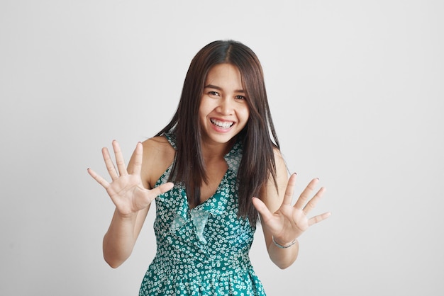 Feliz chica asiática tailandesa con un vestido verde de verano muestra diez dedos y se ríe alegremente aislada en un fondo blanco, pide que se detenga