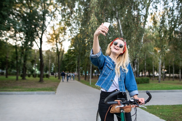 Feliz chica atractiva en el parque con la bicicleta para tomar selfie smartphone y sonrisas.