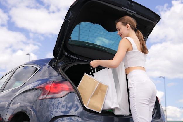 Feliz chica elegante positiva joven hermosa mujer de moda está abriendo el baúl del auto sonriendo en el estacionamiento del centro comercial de compras llevando bolsas de papel de compras en la mano al aire libre Copiar espacio para texto