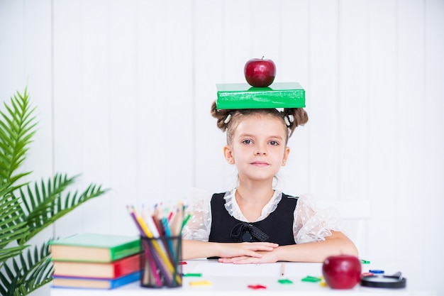 Feliz chica inteligente en uniforme escolar mantenga libro y manzana roja en la cabeza, mire a la cámara.