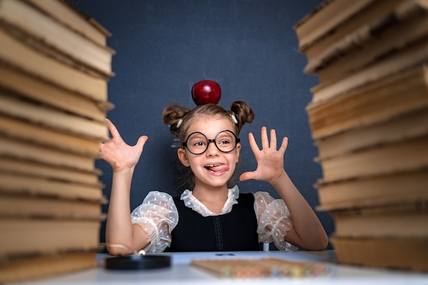 Feliz chica inteligente en vasos redondeados con manzana roja en la cabeza sentada entre dos pilas de libros, divertirse y mirar a la cámara sonriendo.