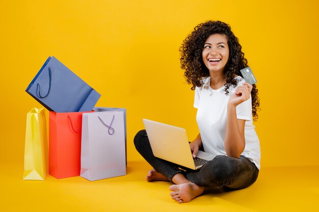 Feliz chica negra con coloridos bolsos de compras sentado con laptop y tarjeta de crédito aislado sobre amarillo