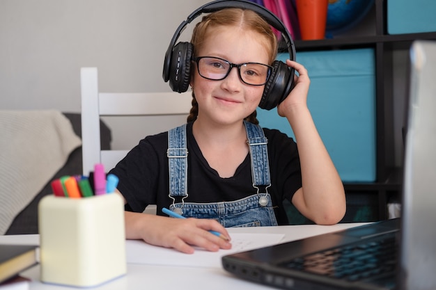 feliz chica pelirroja en anteojos escuchar en auriculares mientras estudia en casa. De vuelta a la escuela