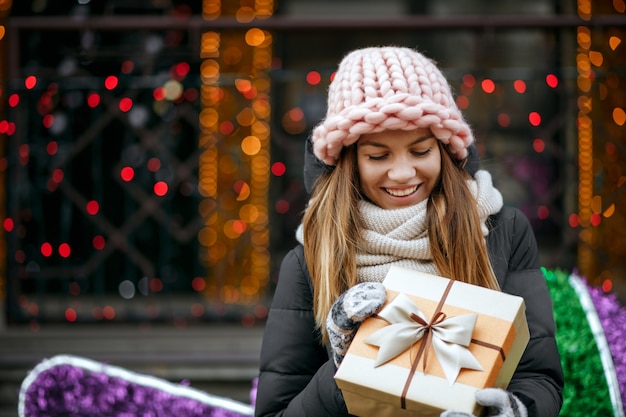 Feliz chica rubia con gorro rosa tejido y bufanda con caja de regalo. Espacio para texto