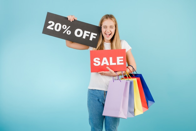 Foto feliz chica rubia grita con 20% de descuento en cartel de venta y coloridas bolsas de compras aisladas sobre azul