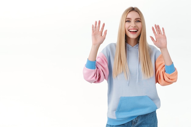 Feliz chica rubia guapa en sudadera con capucha, agitando ambas manos en señal de despedida o hola