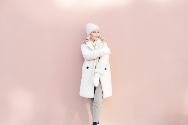 Foto feliz chica rubia en ropa de invierno blanco posando contra una pared de color rosa.