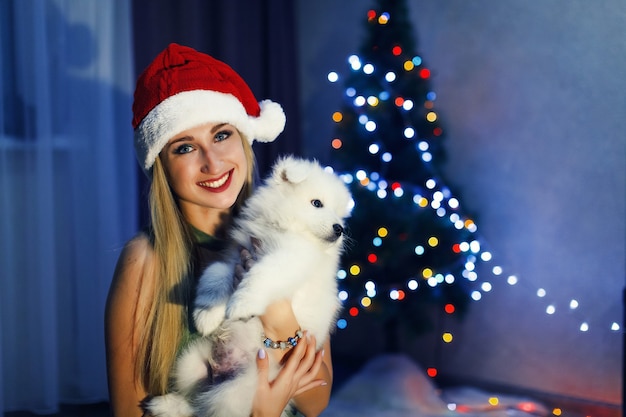 Feliz chica rubia con sombrero de navidad con perro husky samoyedo en adornos navideños