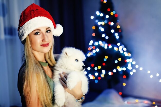 Feliz chica rubia con sombrero de navidad con perro husky samoyedo en adornos navideños