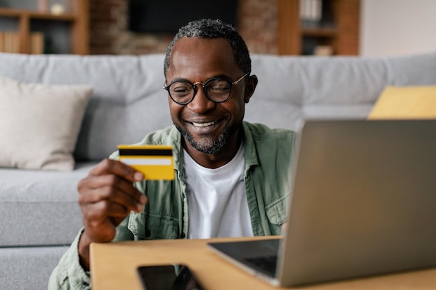 Feliz chico afroamericano de mediana edad con gafas y casual disfruta de las compras en línea en una computadora portátil con tarjeta de crédito