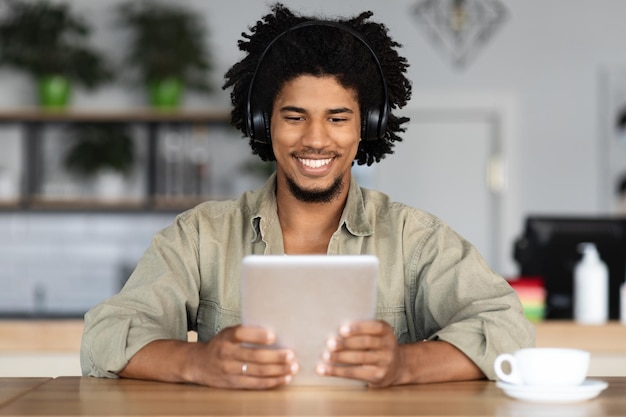 Feliz chico rizado negro milenario en auriculares mirando tablet surfeando en internet sentado en sits