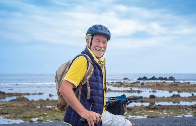 Feliz ciclista barbudo sênior usando capacete e mochila em pé ao longo da praia do mar olhando para a câmera