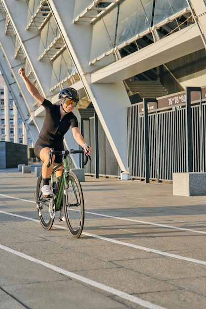 Feliz ciclista profesional en ropa de ciclismo negra y equipo de protección sonriendo elevándola