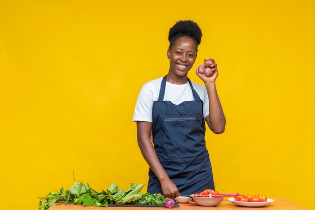 Feliz cocinera africana sosteniendo algunas cebollas con otros ingredientes alimentarios y vegetales frente a ella en un escritorio