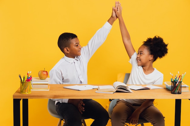 Feliz colegial afro-americana e estudante de raça mista sentados juntos na mesa e dando mais cinco um ao outro em fundo amarelo De volta ao conceito de escola