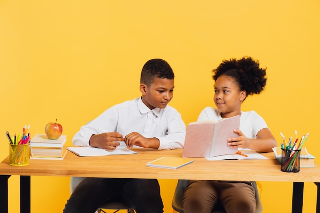 Feliz colegial afro-americana e estudante de raça mista sentados juntos na mesa e estudando em fundo amarelo de volta ao conceito de escola