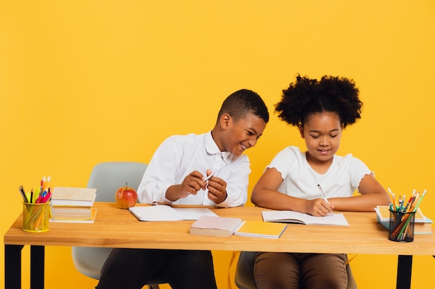 Feliz colegial afro-americana e estudante de raça mista sentados juntos na mesa e estudando em fundo amarelo de volta ao conceito de escola