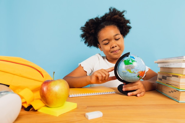 Feliz colegial afro-americana sentada na mesa na sala de aula e olhando para o globo do mapa do mundo em fundo azul De volta ao conceito de escola