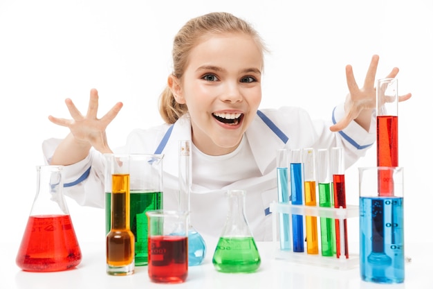 Feliz colegial com jaleco branco fazendo experimentos químicos com líquido multicolorido em tubos de ensaio isolados sobre uma parede branca