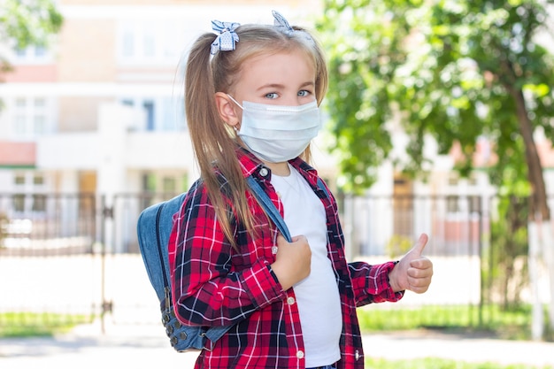 Feliz colegial em uma máscara protetora com uma mochila mostra como. em uma camiseta branca e uma camisa xadrez