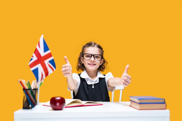 Feliz colegiala caucásica sentado en el escritorio mostrando Thumbs up lección de inglés bandera de gran bretaña