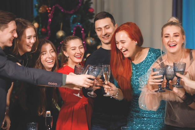 Feliz Companhia de amigos celebra o Natal ou o Ano Novo em uma mesa perto da árvore de Natal com guirlandas. Mulheres riem, tilintam taças com champanhe.