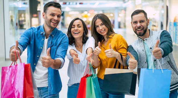 Feliz compañía de sonrientes amigos hermosos emocionados con estilo con bolsas de papel de colores se divierten y muestran los pulgares hacia arriba mientras compran en el centro comercial.
