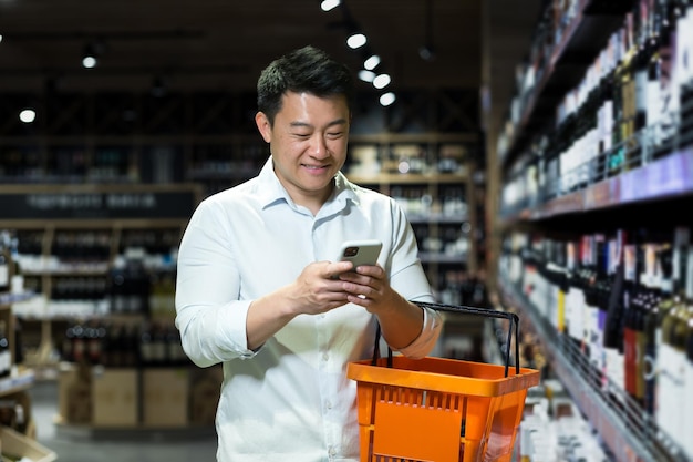 Feliz comprador asiático no supermercado usando aplicativo de smartphone para selecionar e escanear vinho na loja entre