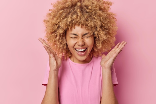 Feliz concepto de emociones positivas Una joven alegre con el pelo tupido cury mantiene las palmas levantadas exclama en voz alta recapitula sobre noticias increíbles vestida con una camiseta informal aislada sobre un fondo rosa