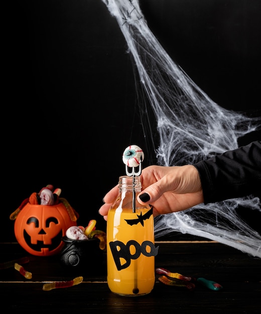 Feliz concepto de Halloween. Mano de mujer con uñas negras sosteniendo cóctel de Halloween colorido aterrador con decoraciones de fiesta sobre fondo oscuro