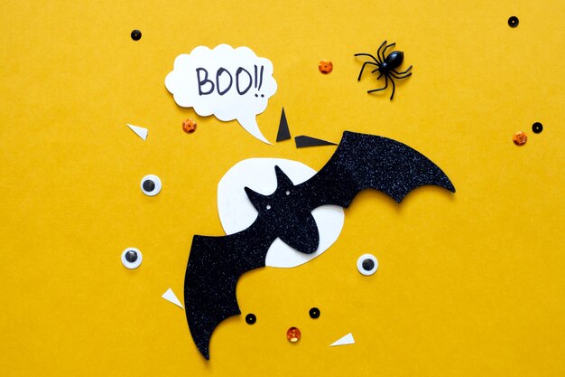 Feliz concepto de vacaciones de halloween. Murciélagos de papel brillo negro y luna sobre fondo amarillo brillante con araña negra, ojos, confeti. Tarjeta de felicitación de fiesta de Halloween. Ortografía de la palabra Boo.