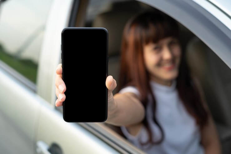 Feliz conductora sonriendo y mostrando el teléfono inteligente mientras está a través de la ventana del auto