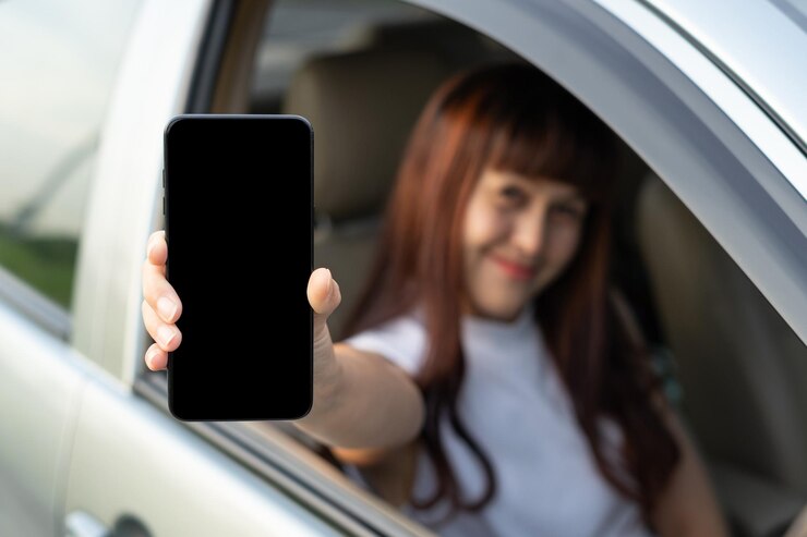 Feliz conductora sonriendo y mostrando el teléfono inteligente mientras está a través de la ventana del auto