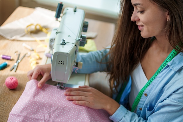 Feliz costurera sonriente con máquina de coser eléctrica y diferentes accesorios de costura durante el proceso de confección en el lugar de trabajo