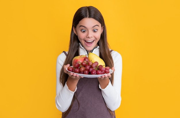 Feliz criança espantada segura prato de frutas frescas em vitaminas de fundo amarelo