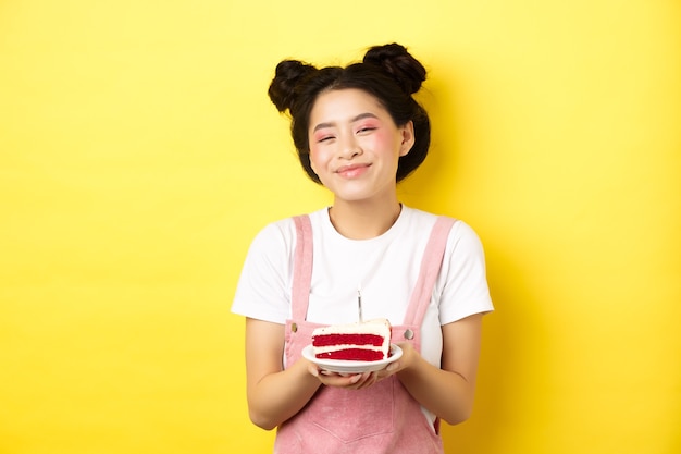 Feliz cumpleañera asiática con maquillaje brillante, soplar velas en la torta, pedir deseos, de pie en amarillo.