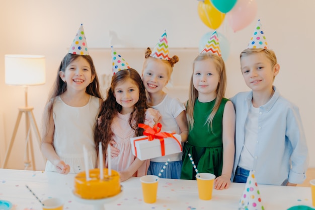 Feliz cumpleañera se encuentra con presenet, amigos felices vienen a felicitarla, usan sombreros de cono de fiesta, se paran cerca de la mesa festiva con pastel