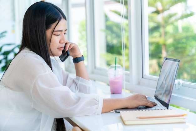 Feliz da mulher de negócios de freelancers asiáticos falando fazendo telefonema no smartphone casual trabalhando com computador portátil para navegar na internet conversando e blogando no café