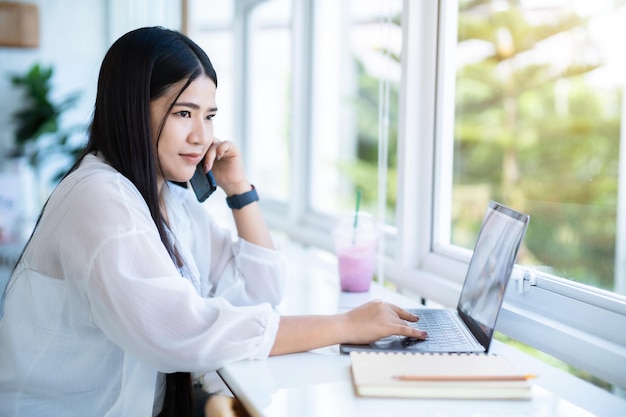 Feliz da mulher de negócios de freelancers asiáticos falando usando smartphone casual trabalhando com laptop com notebook para navegar na internet conversando e blogando em um café público