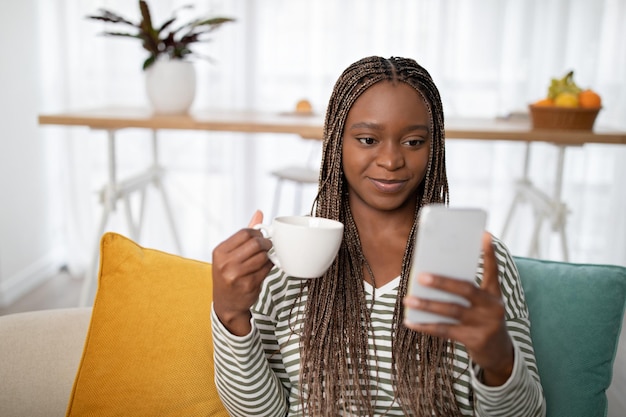 Feliz dama africana sentada en el sofá con un teléfono inteligente tomando café