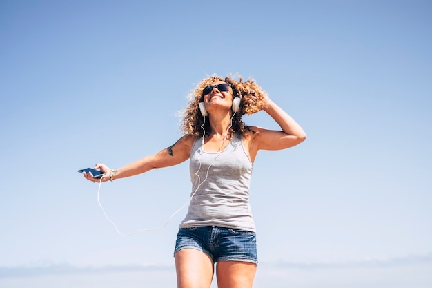 Feliz dama alegre con cabello rizado rubio escuchando música con auriculares y teléfono móvil al aire libre divirtiéndose y disfrutando de la vida